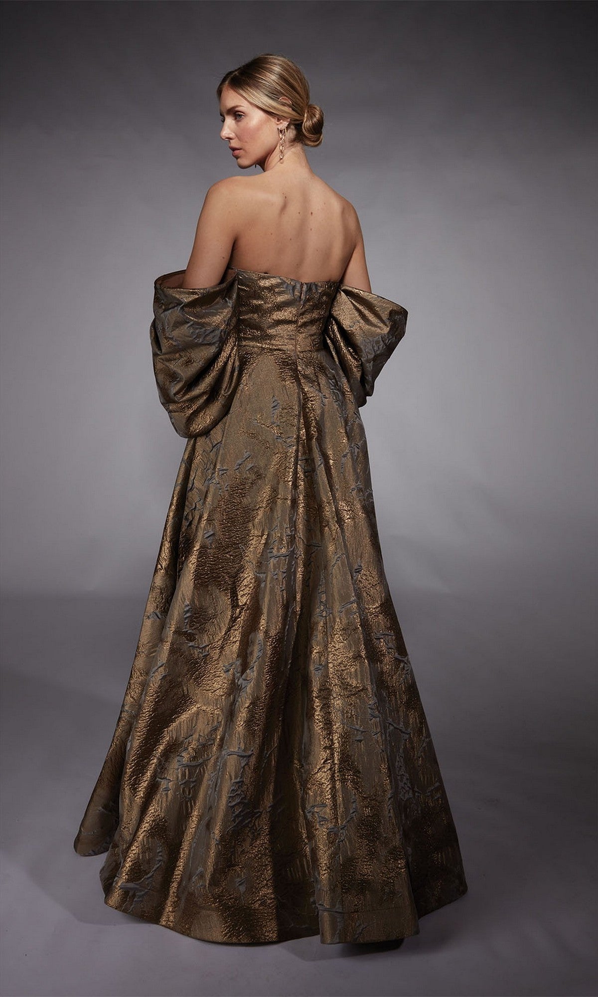 Wearing the Alyce 27760 long formal dress with detachable puff sleeves, a woman faces away, her hair in a low bun, highlighting the elegant jacquard metallic fabric against a neutral backdrop.